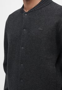 Cardigan en tricot gris foncé à boutons avec col côtelé, poche latérale et logo de marque brodé discret sur la poitrine.