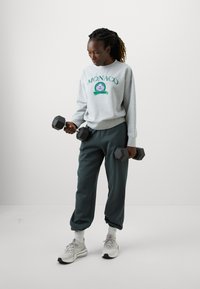 Šedá mikina s textom "MONACO", tmavozelené tepláky a šedé tenisky. Drží čierne hexagonálne činky. Jednoduché pozadie.