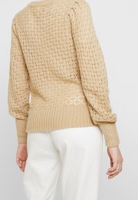 Pull beige en maille avec un motif texturé et un ourlet côtelé. Manches longues à coupe ample, en matériau léger et respirant.