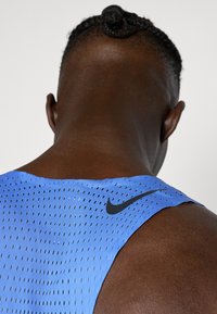 Niebieski sportowy top na ramiączkach z siateczkową teksturą, z czarnym logo Nike na tylnej części ramienia. Gładki materiał, bez rękawów.