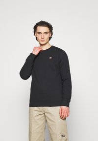 Hanorac negru cu manșete și tiv cu nervuri, având un mic logo brodat pe piept. Purtați cu pantaloni ușor bej.