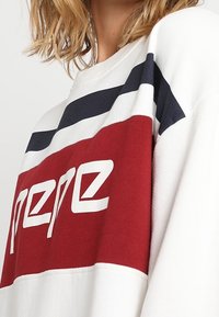 Felpa bianca a maniche lunghe con strisce orizzontali rosse e blu navy; presenta un logo testuale bianco in evidenza sulla sezione rossa, con dettagli di cucitura visibili.