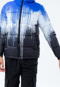 Giacca imbottita con un design a gradiente che passa dal blu al bianco e al nero, con motivi di gocce di vernice. Chiusura con zip e tasche laterali.