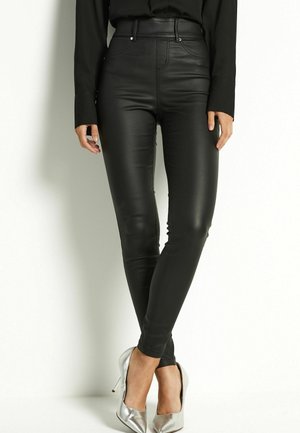 Femme portant des leggings noirs en simili cuir taille haute et des chaussures à talons hauts argentées, debout sur un fond blanc.