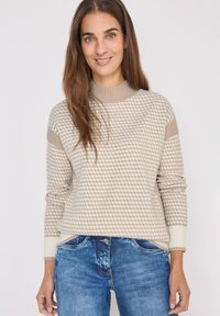 Beżowy sweter z wzorem, wykonany na drutach, o wysokim dekolcie i ściągaczach przy rękawach, w zestawieniu z niebieskimi dżinsami. Sweter ma chropowaty, geometryczny wzór.