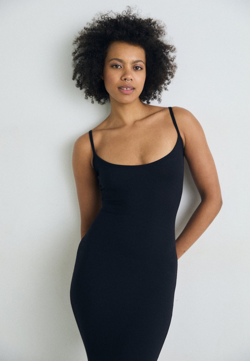 Robe midi noire côtelée avec des fines bretelles spaghetti et un col rond, présentant une silhouette ajustée qui met en valeur la forme du corps.