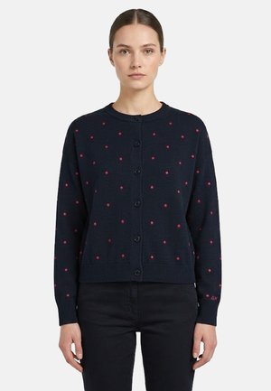 Donna che indossa un cardigan blu navy con bottoni e pois rosa, abbinato a pantaloni neri, in piedi davanti a uno sfondo semplice.