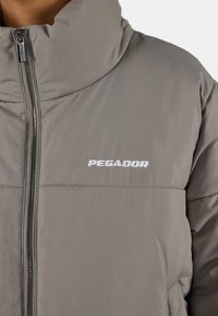 Grå polstret jakke med høy krage, med frontglidelås og "PEGADOR"-logo i hvit brodering på brystet. Glatt tekstur.