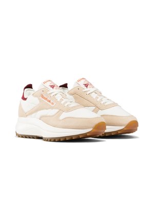 Bílo-béžové tenisky Reebok s gumovou podrážkou, šněrováním vpředu a červeným logem na boku a jazyku, zobrazené mírně šikmo.