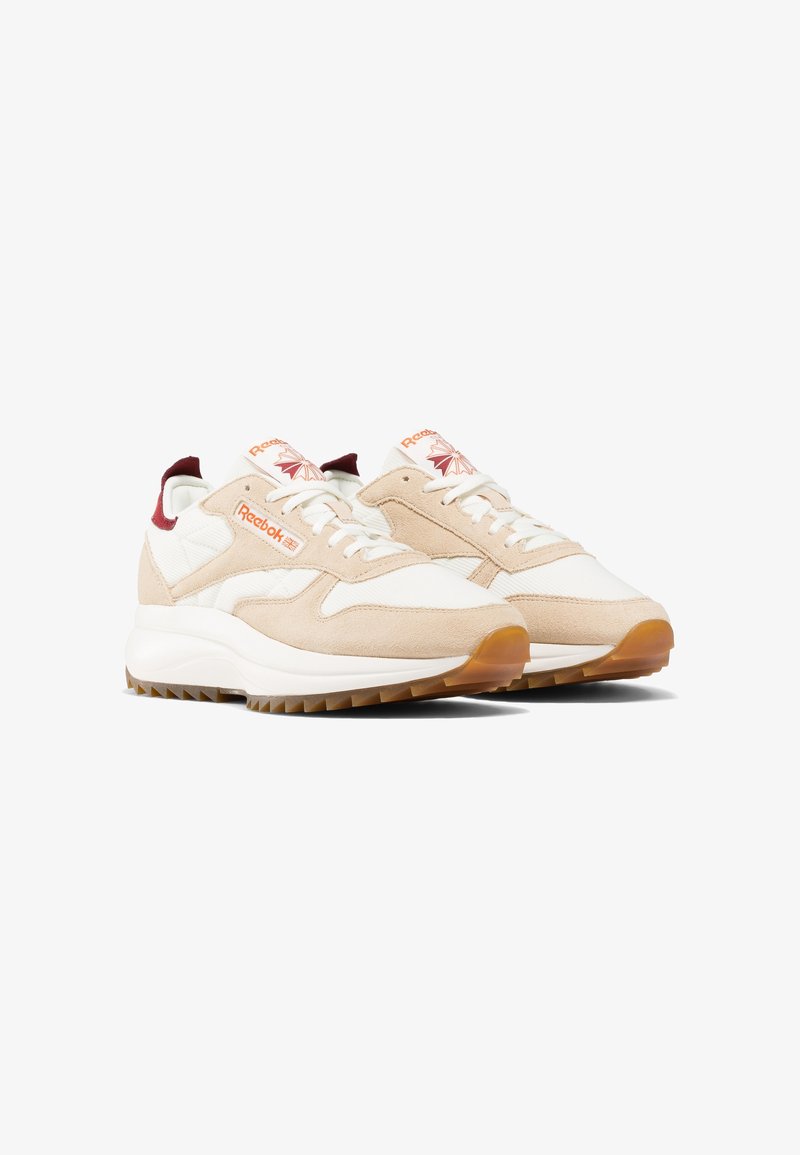 Baskets blanches et beiges Reebok avec semelles en gomme, devant à lacets et logo rouge sur le côté et la languette, présentées sous un léger angle.