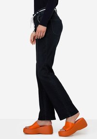 Vaqueros oscuros de cintura alta con un cinturón de cordón blanco, combinados con sandalias de plataforma de un brillante color naranja, que presentan una textura suave y una forma redondeada.