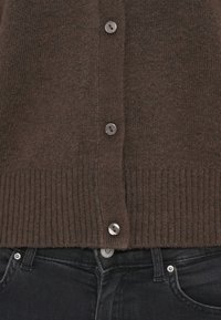 Bruine gebreide cardigan met geribde zoom en vijf metalen knopen. Zwarte jeans zichtbaar aan de onderkant, met een klein knoopdetail.