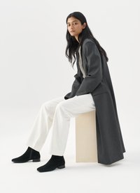 Šedý tailored kabát s kontrastní vnitřní podšívkou, bílé široké kalhoty a černé semišové kotníkové boty. Model sedící na světlém kostce.