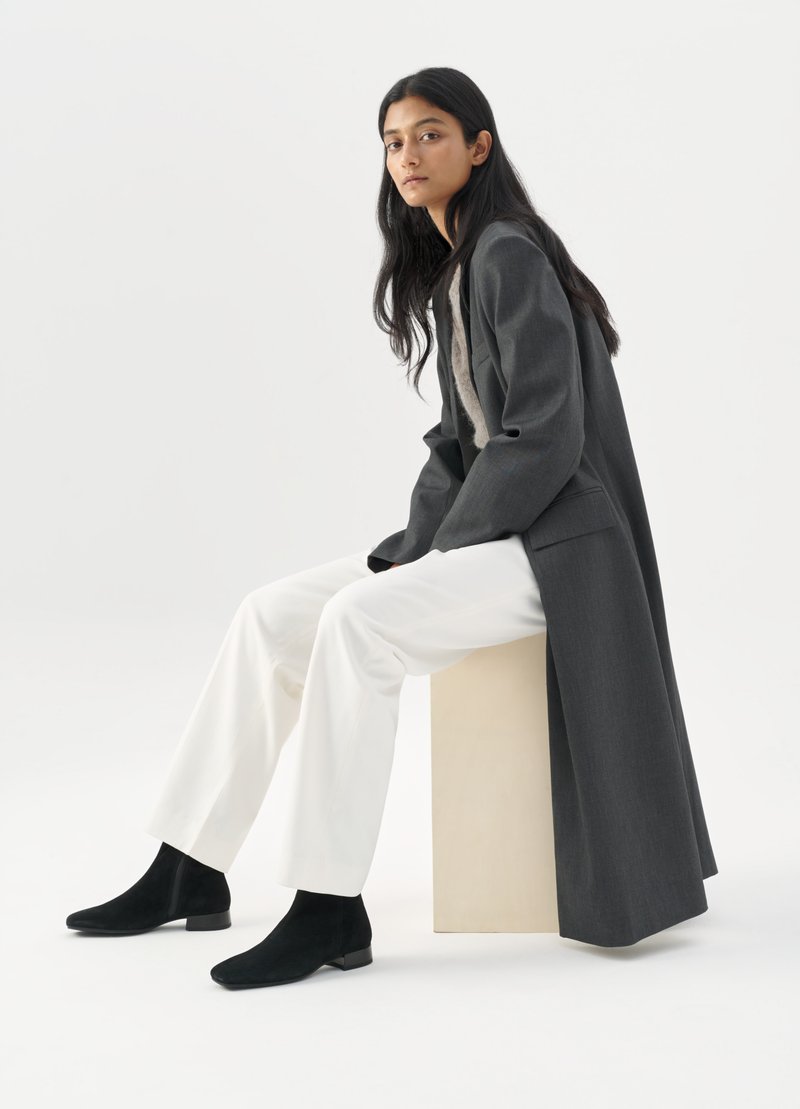 Grau taillierter Mantel mit kontrastierendes Innenfutter, weiße weite Hose und schwarze Wildleder-Ankle Boots. Modell sitzt auf einem hellen Würfel.