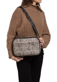 Borsa a spalla in canvas beige con motivi a logo neri, chiusura con zip e tracolla regolabile con branding. Forma rettangolare.