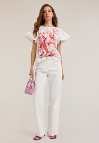Top bianco con design floreale rosa, maniche a volant e pantaloni bianchi a gamba larga. Tiene in mano una piccola borsa rosa e indossa sandali con tacco rosa.
