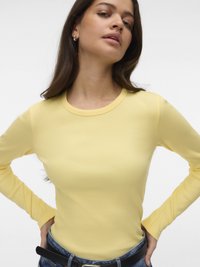 Kvinne med langt brunt hår iført en gul ribbestrikket langermet skjorte, blå jeans og hoops-øreringer, poserer med hendene på hoftene.