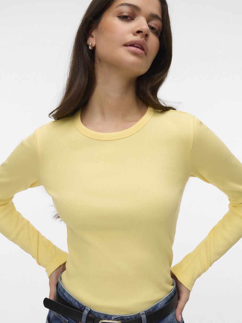 Kvinne med langt brunt hår iført en gul ribbestrikket langermet skjorte, blå jeans og hoops-øreringer, poserer med hendene på hoftene.