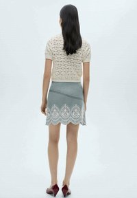 Pull beige à manches courtes en tricot avec un motif en dentelle, associé à une jupe grise avec un ourlet festonné et des détails en dentelle. Chaussures rouge foncé.