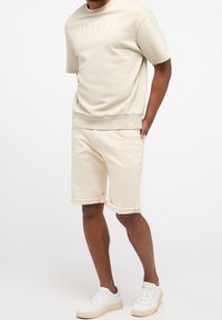 Sweat-shirt beige avec logo "MUSTANG", manches courtes, et short beige assorti à revers, associé à des baskets blanches.