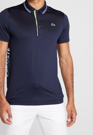 Vue recadrée d’un modèle portant un polo bleu marine à col rayé blanc, avec une bordure vert citron sur la patte de boutonnage, un logo crocodile et un pantalon blanc.