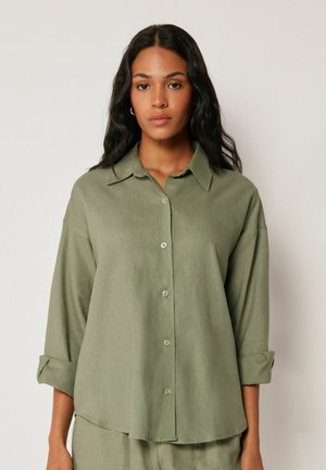 Kvinde med langt mørkt hår iført en oversized olivengrøn skjorte med knapper og opkrogede ærmer, stående mod en ensfarvet lys baggrund.
