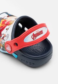 Crocs sur le thème des Avengers, avec une tige à motif bleu et blanc, une sangle rouge avec logo et une semelle en caoutchouc noir avec un grip texturé.