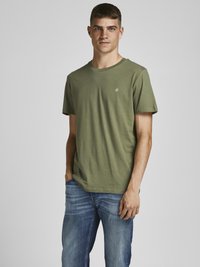 Camiseta de algodón color verde oliva, de cuello redondo y manga corta, con un pequeño logo blanco en el pecho; combinada con jeans de mezclilla azul.