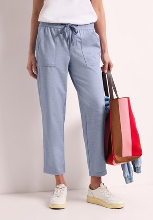 Person iført lyseblå snorebukser og hvide sneakers, der holder en stor stribet taske med pink, rød og brun farve.