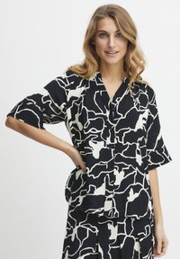 Sort-hvid mønstrede kortærmede bluse med krave, der har abstrakte designs og knaplukning foran. Letvægtsstof.