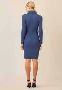 Abito maglione blu a costine con colletto alto, maniche lunghe, forma aderente e lunghezza fino al ginocchio. Indossato con scarpe con tacco nere e cinturini.