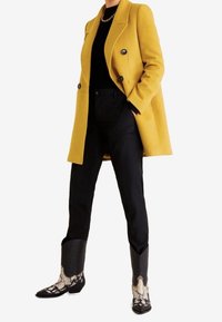 Geel mantel met dubbele rij knopen, zwart topje en broek, zwarte cowboylaarzen met witte slangprintdetails. Overal gladde texturen.