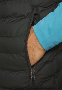 Gilet rembourré noir avec une poche à main zippée sur le côté gauche, dotée d'une fermeture éclair métallique. L'arrière-plan comprend une chemise à manches longues bleue.