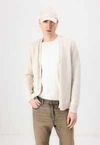 Light beige knitted cardigan with buttons, worn over a white t-shirt, paired with light brown jeans and a matching cap, set against a white background.