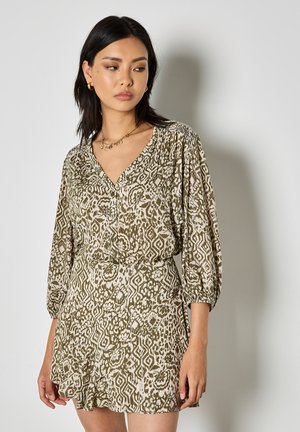 Femme portant une robe à col en V beige et vert olive à motifs avec des manches trois-quarts, des boucles d'oreilles créoles dorées et un collier en or sur un fond uni.