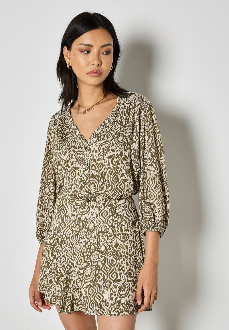 Femme portant une robe à col en V beige et vert olive à motifs avec des manches trois-quarts, des boucles d'oreilles créoles dorées et un collier en or sur un fond uni.