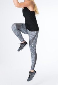 Černý sportovní tílko spárované s vzorovanými šedými legínami s geometrickými designy a akcenty. Obuv ve šedé barvě s černými šňůrkami.