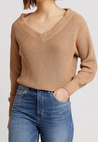 Beige knitted sweater with a V-neck, long sleeves, and ribbed texture, paired with blue high-waisted jeans.