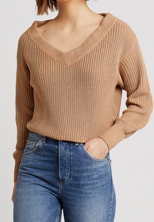 Maglione lavorato a maglia beige con scollo a V, maniche lunghe e texture a costine, abbinato a jeans blu a vita alta.