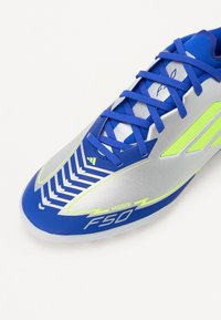 Blauw en zilveren voetbalschoen met felgroene accenten, voorzien van een gestructureerde bovenkant, slank ontwerp en opvallende "F50" en "Messi" branding.