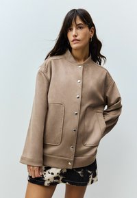 Jachetă din suede maro cu guler rotund, închidere cu capse și buzunare frontale. Modelul poartă o fustă cu un model alb și negru.