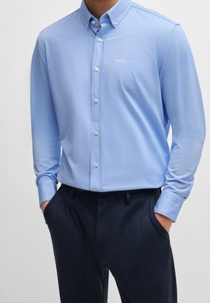 Mann trägt ein hellblaues Hemd mit Knöpfen und "BOSS"-Logo auf der Brust sowie dunkelblaue Hosen, Hände in den Taschen, steht vor einfachem Hintergrund.