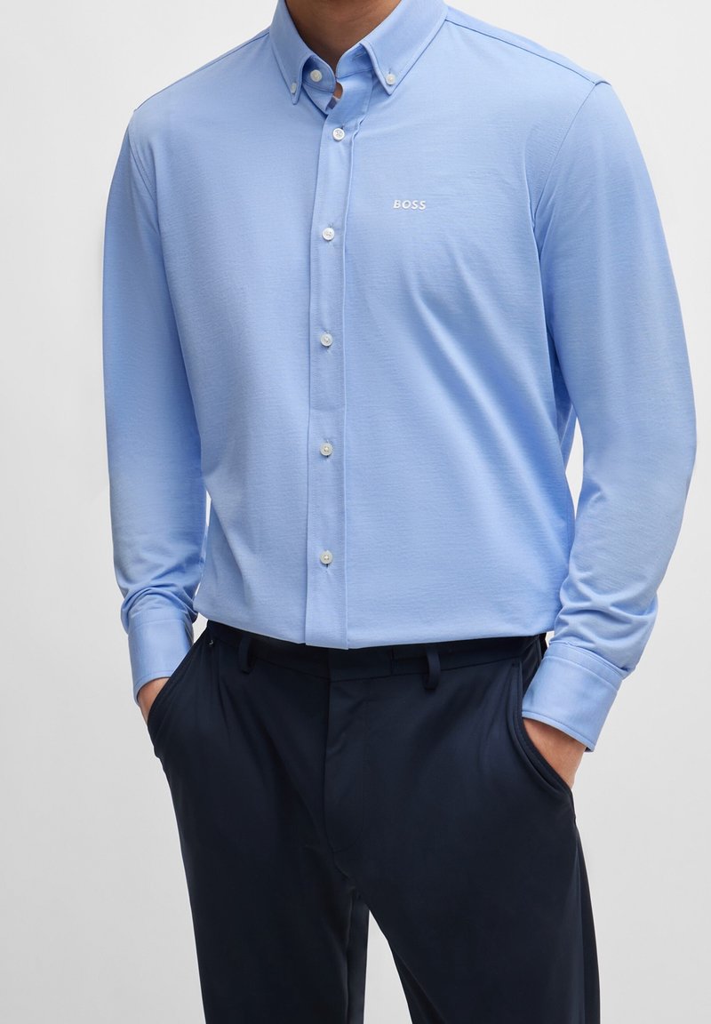 Mand iført en lyseblå skjorte med knapper og "BOSS"-logo på brystet samt mørkeblå bukser, hænderne i lommerne, stående mod en ensfarvet baggrund.