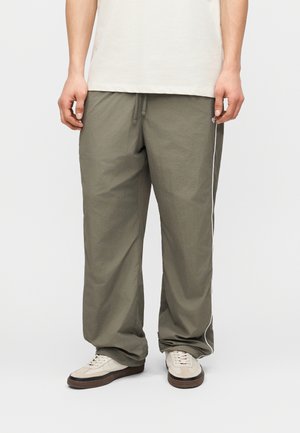 Oseba v širokih olivno zelenih hlačah z belim bočnim črtom, bež supergah in kremno obarvani srajci stoji na enobarvnem ozadju.