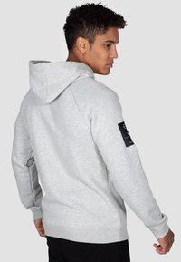 Light gray hoodie featuring a kangaroo pocket, ribbed cuffs, and hem, with a black fabric patch on the left sleeve.