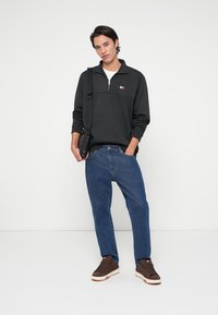 Σκούρο γκρι πουλόβερ με φερμουάρ μέχρι το μισό και τσέπη, σε συνδυασμό με μπλε τζην και καφέ αθλητικά παπούτσια. Η τσάντα είναι μαύρη και φορεμένη χιαστί.