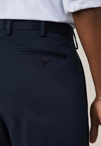 Pantaloni blu navy realizzati in tessuto liscio, dotati di una tasca posteriore con chiusura a bottone e un design slim e affusolato.