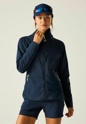 Woman wearing navy outdoor jacket and shorts, adjusting jacket zipper, with blue cap and reflective sunglasses on cap, standing against plain background.