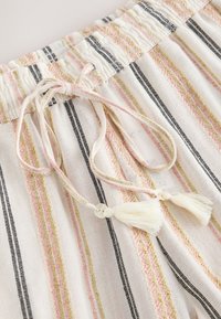 Tissu blanc avec des rayures verticales noires, roses et beige, doté d'une taille élastique, d'un lien coulissant et de pompons aux extrémités.