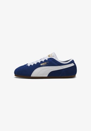 Blauwe suède Puma-sneaker met witte streep, witte veters en gouden Puma-logo aan de zijkant en op de tong, gezien van de zijkant.